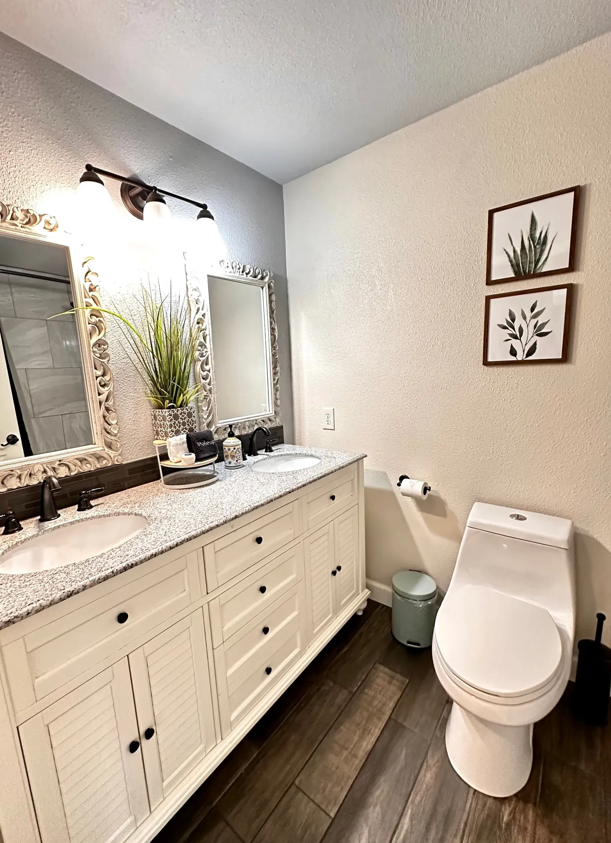 This image features a bright, modern bathroom with dual sinks, a large decorative mirror, and framed botanical prints. A potted plant adds a touch of freshness, and the overall atmosphere is tranquil and inviting, suggesting a well-kept, shared amenity space.