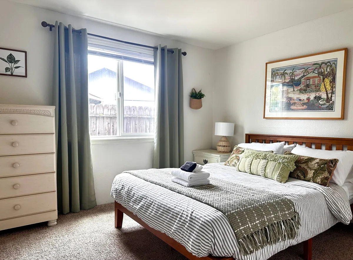 A cozy bedroom scene with a plush bed, soft curtains that let in morning light, a dresser, and tranquil artwork. The atmosphere is calm and restful, ideal for unwinding after a busy day.
