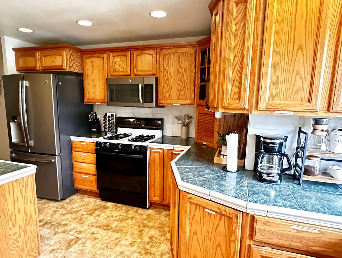 This image shows a welcoming kitchen with a stainless steel refrigerator, black gas stove, warm wooden cabinetry, and blue-green countertops. The setting is spacious, practical, and ideal for daily routines like cooking or gathering with family.