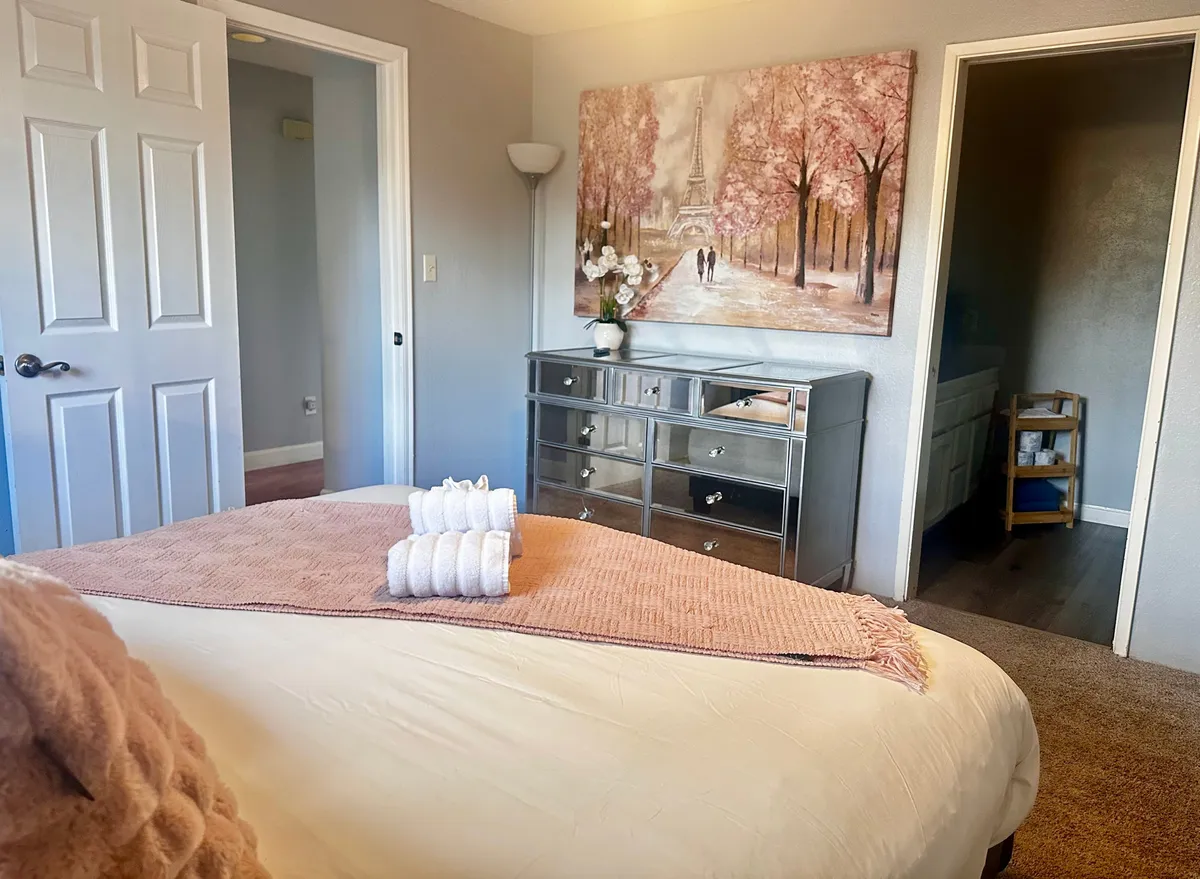A softly lit bedroom with a comfortable bed made up in plush linens, decorative towels placed neatly at the foot, and Paris-themed artwork above a dressing table. Light filters through the window, creating a serene and inviting space.