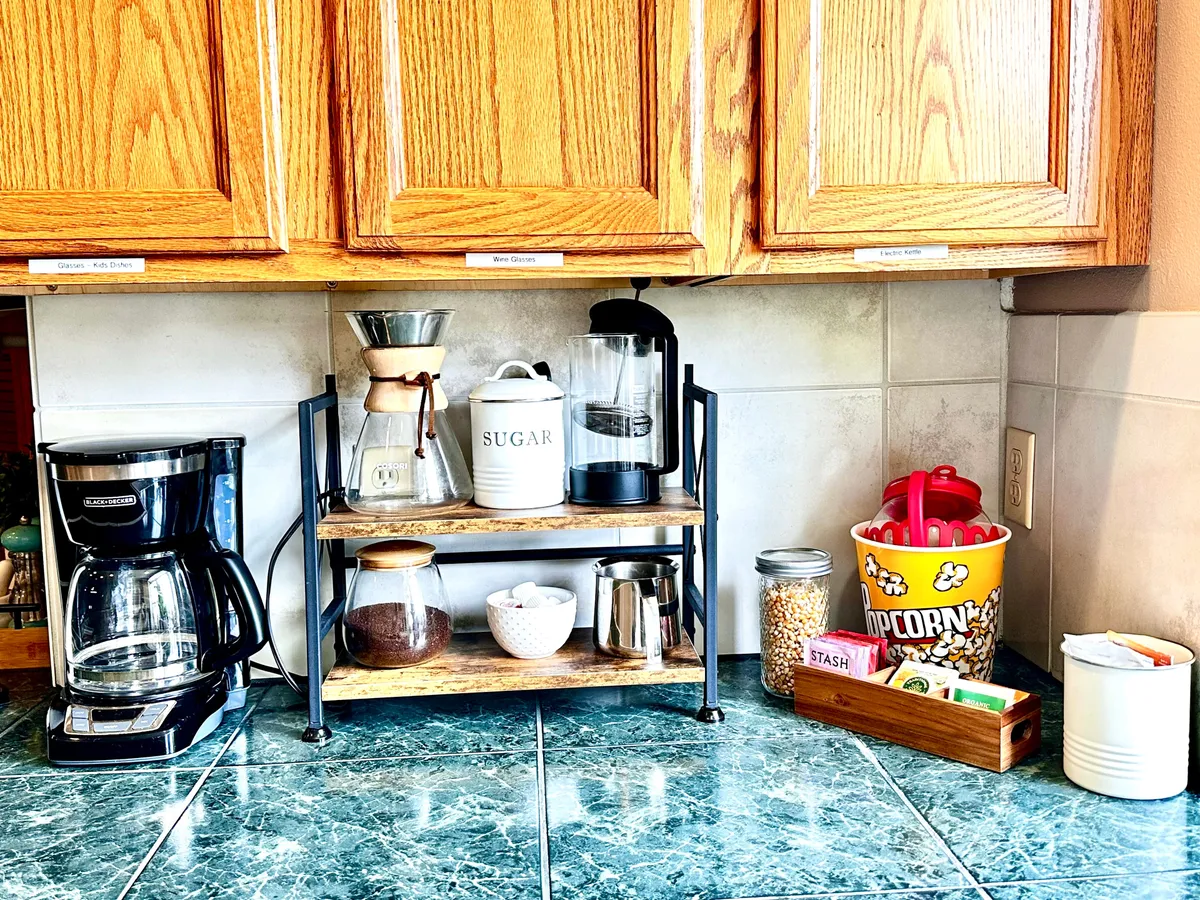 The image highlights a cozy kitchen area with a coffee maker, grinder, sugar container, and a popcorn bucket displayed on shelves with jars. The countertop invites families to gather for morning coffee or snacks, while the popcorn bucket hints at movie nights and shared activities.