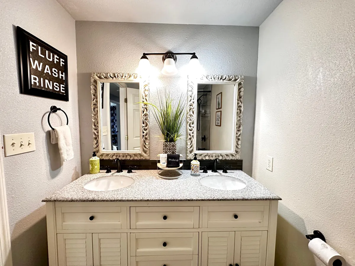 A stylish, newly remodeled bathroom featuring a double vanity with sinks, ornate mirrors, a decorative sign, and lush potted plant atop a granite countertop. The space feels welcoming and ideal for self-care routines.