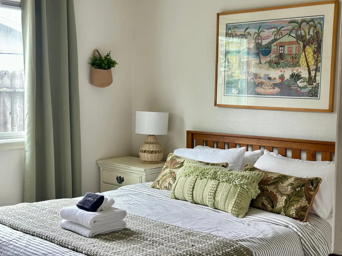 A serene bedroom scene showcasing a bed with decorative pillows, framed artwork above the headboard, a woven lamp, and a nightstand with a plant. Sunlight gently filters in, enhancing the tranquil and rejuvenating feel of the space.